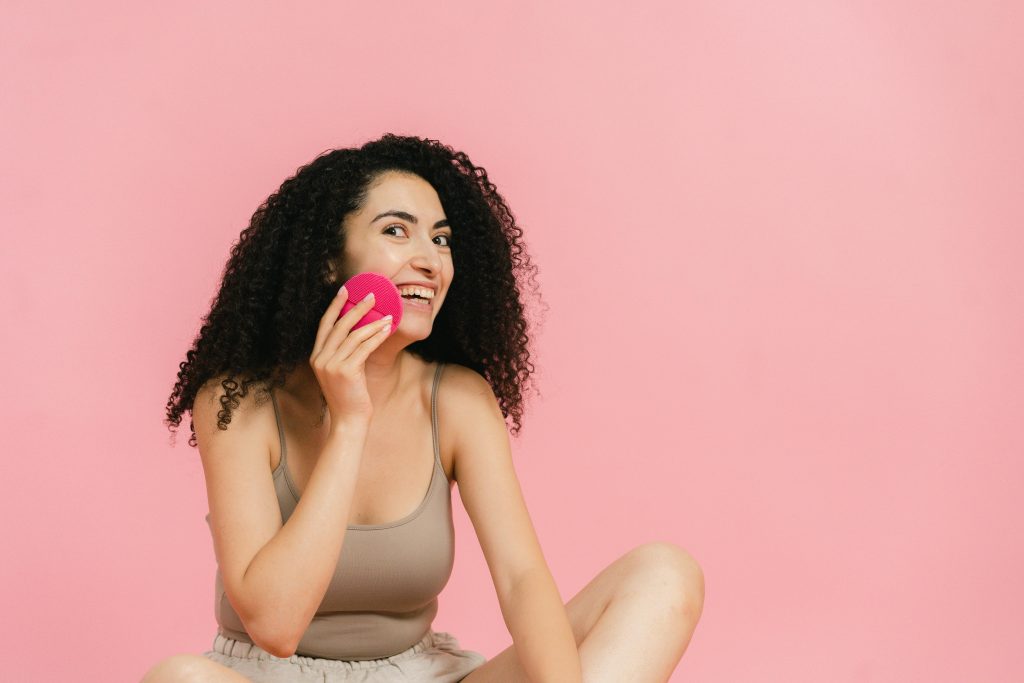 A cheerful young woman with curly hair enjoys her skincare routine against a pink backdrop. Studio shot.